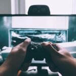 A person holds a video game controller in front of a TV